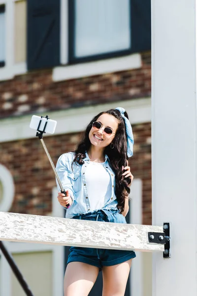 Attractive woman holding selfie stick and taking selfie on smartphone — Stock Photo