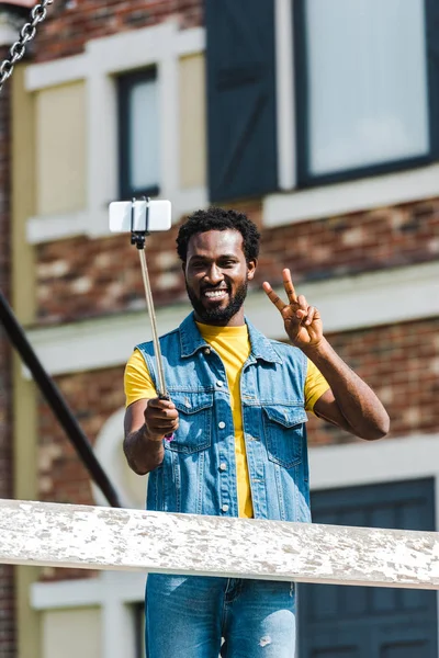 Hombre afroamericano feliz mostrando signo de paz mientras sostiene selfie palo y hablar selfie - foto de stock