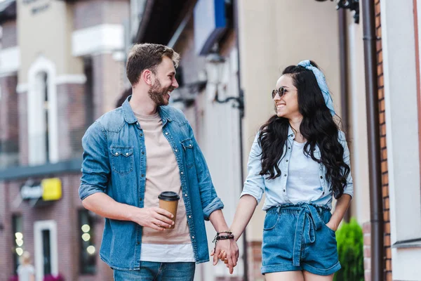 Cheerful woman with hand in pocket walking and holding hands with man — Stock Photo