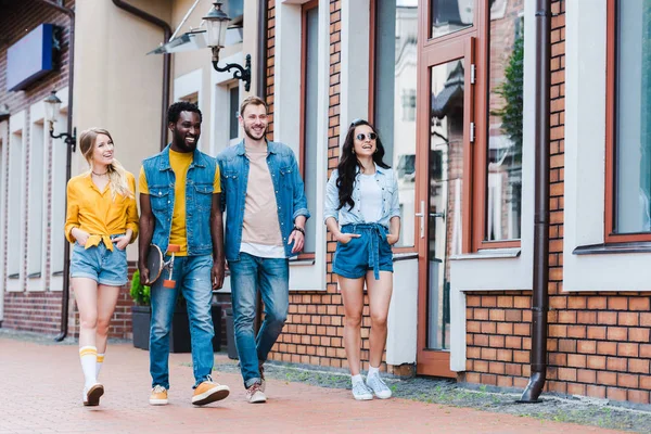 Chicas felices con las manos en los bolsillos caminando con amigos alegres multiculturales - foto de stock