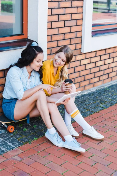 Femme joyeuse pointant avec le doigt à la tablette numérique tout en étant assis sur le panneau de penny avec un ami — Photo de stock