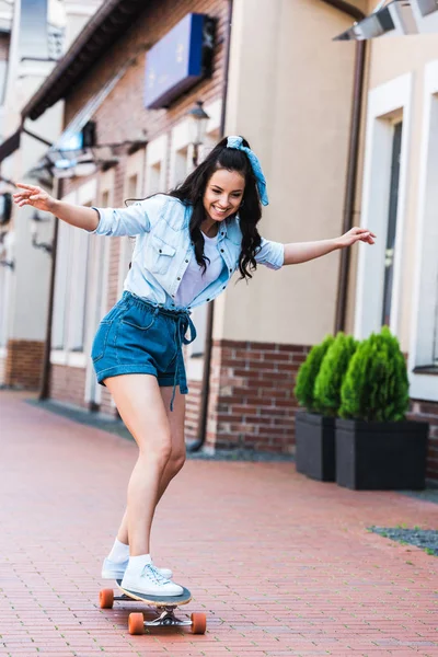 Happy young woman with outstretched hands riding penny board near building — Stock Photo