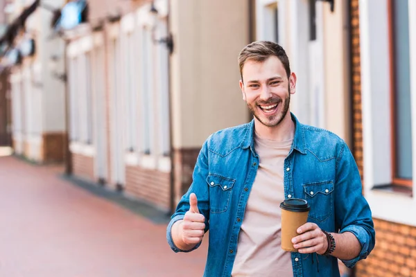 Joyeux bel homme tenant tasse en papier et souriant tout en montrant pouce vers le haut — Photo de stock