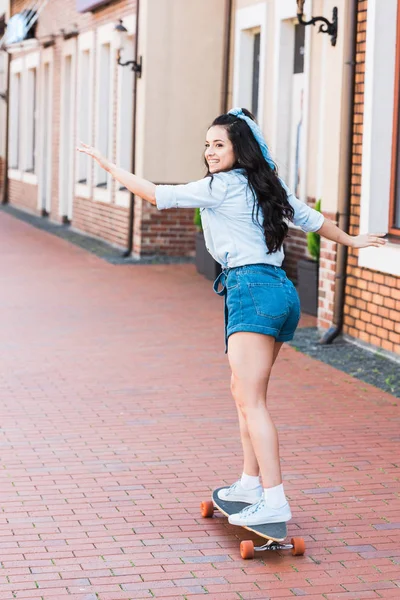 Cheerful young woman with outstretched hands riding penny board near building — Stock Photo