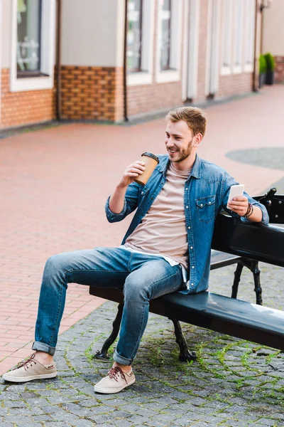 Hombre feliz sosteniendo la taza de papel y el teléfono inteligente mientras está sentado en el banco - foto de stock