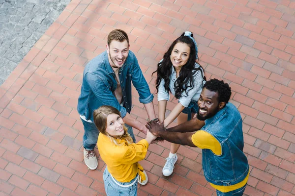 Overhead view of cheerful multicultural friends putting hands together and looking at camera — Stock Photo