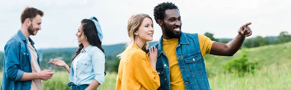 Panoramaaufnahme eines glücklichen afrikanisch-amerikanischen Mannes, der mit dem Finger zeigt, während er mit einem Mädchen in der Nähe von Freunden steht — Stockfoto