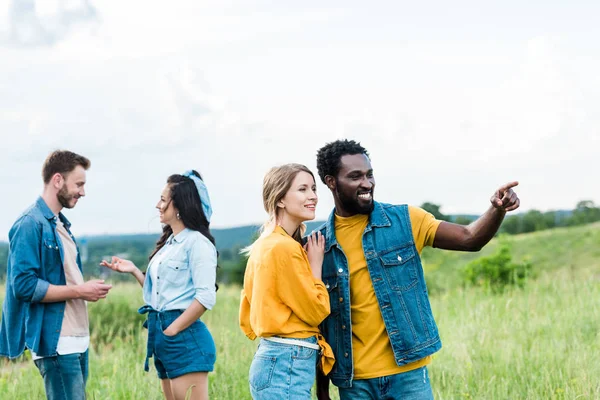 Hombre afroamericano feliz señalando con el dedo mientras está de pie con la chica cerca de amigos - foto de stock