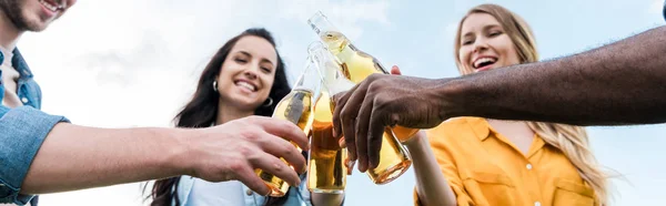 Plano panorámico de alegre multicultural hombres y mujeres tintineo botellas con cerveza - foto de stock