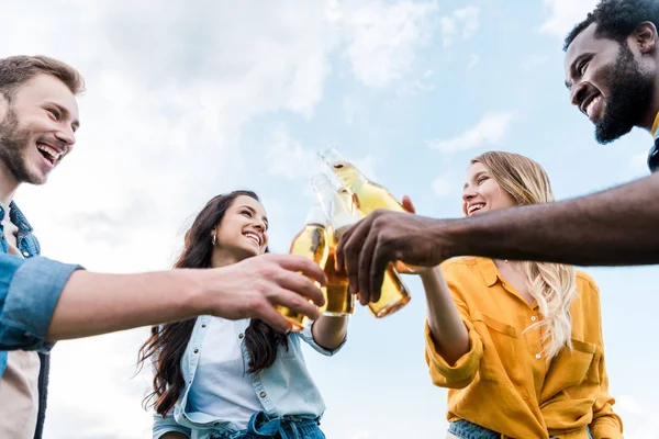 Punto de vista de bajo ángulo de alegre multicultural mujeres y hombres tintineo botellas con cerveza - foto de stock