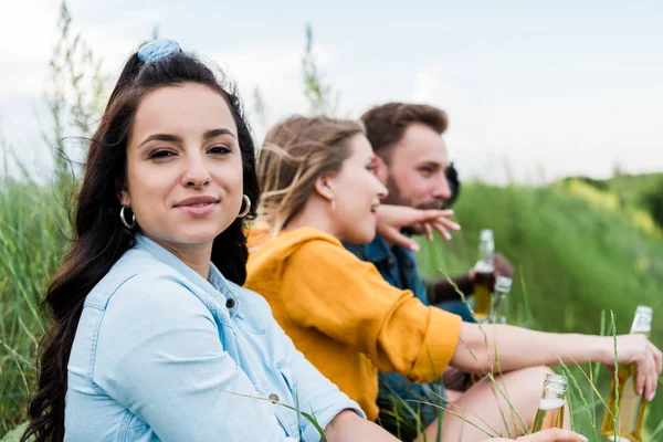 Messa a fuoco selettiva di attraente ragazza guardando fotocamera vicino amici in possesso di bottiglie con birra — Foto stock