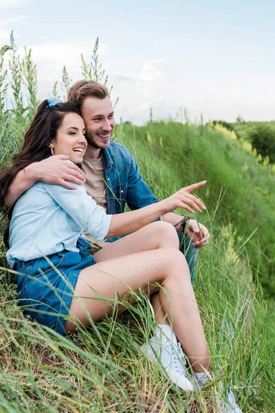 Foco seletivo de alegre jovem mulher sentada com homem bonito na grama verde e apontando com o dedo — Fotografia de Stock