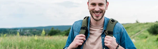 Plan panoramique de jeune homme heureux debout avec sac à dos et regardant la caméra — Photo de stock