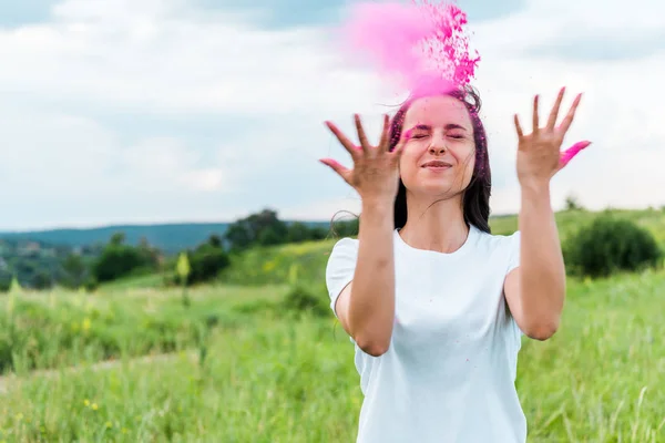 Счастливая молодая женщина стоит с закрытыми глазами и бросает в воздух розовый порошок — стоковое фото