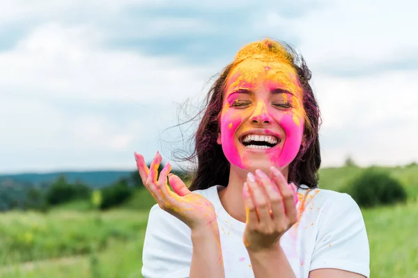 Mujer feliz de pie con los ojos cerrados y rosa y amarillo holi pintura en la cara - foto de stock