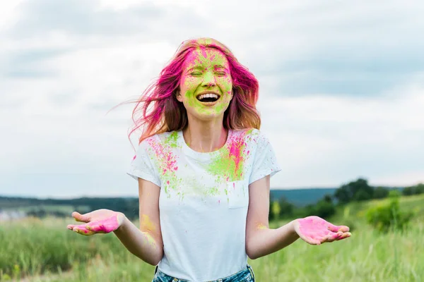 Femme gaie avec les yeux fermés et rose holi peinture sur le visage gestuelle et souriante à l'extérieur — Photo de stock