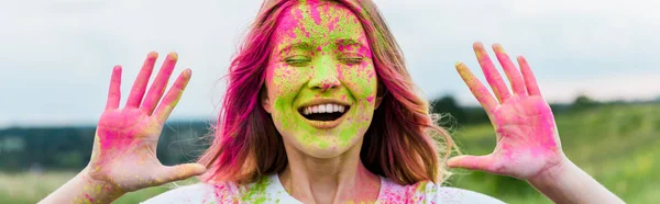 Panoramic shot of cheerful woman with closed eyes and pink holi paint on hands gesturing and smiling outdoors — Stock Photo