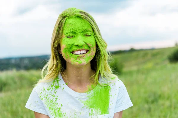 Fröhliche Frau mit geschlossenen Augen und grüner Holi-Farbe im Gesicht, die draußen lächelt — Stockfoto