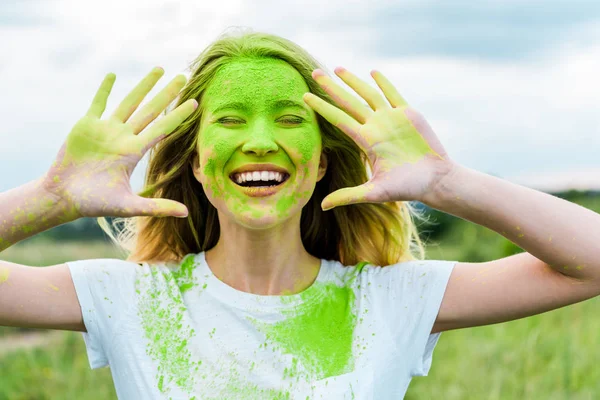 Fröhliche Frau mit geschlossenen Augen und grüner Holi-Farbe auf Händen, die gestikuliert und im Freien lächelt — Stockfoto