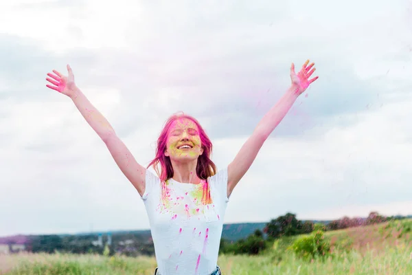 Happy woman with closed eyes and pink holi paint on outstretched hands smiling outdoors — Stock Photo