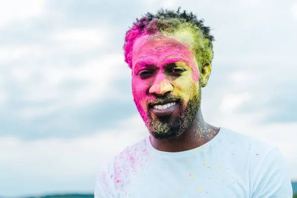 Guapo afroamericano hombre en camiseta blanca y coloridas pinturas holi en la cara - foto de stock