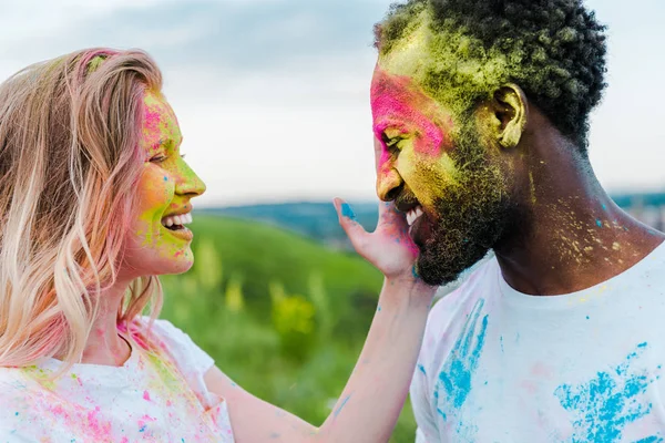 Mujer feliz tocando la cara de hombre afroamericano con pintura holi en la cara - foto de stock