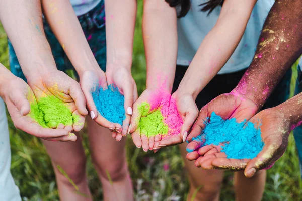 Vista recortada de amigos multiculturales sosteniendo coloridas pinturas holi en las manos - foto de stock