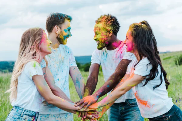 Chicas felices y hombres multiculturales con coloridas pinturas holi en las caras poniendo manos juntas - foto de stock