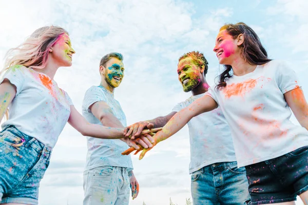 Low angle view of happy multicultural friends with colorful holi paints on faces putting hands together — Stock Photo