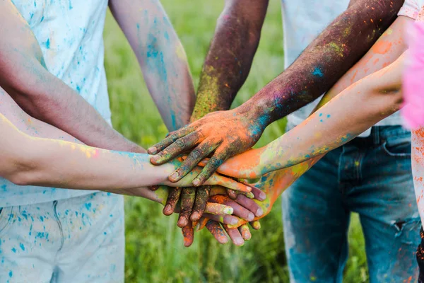 Vista recortada de amigos multiculturales con coloridas pinturas holi poniendo manos juntas - foto de stock