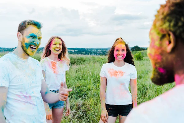 Selective focus of happy man with holi paints on face pointing with finger at african american friend near girls — Stock Photo