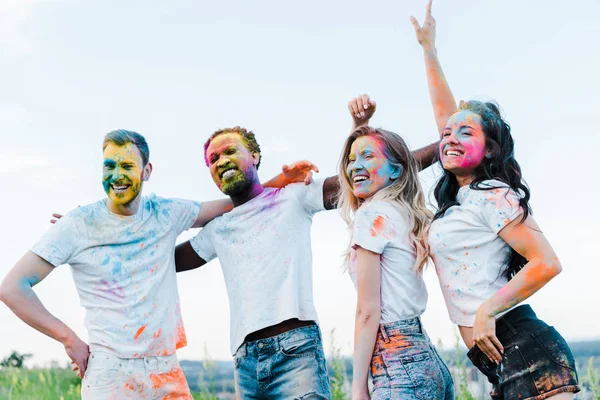 Glücklicher Mann mit Hand auf der Hüfte in der Nähe multikultureller Freunde mit Holi-Farben im Gesicht — Stockfoto