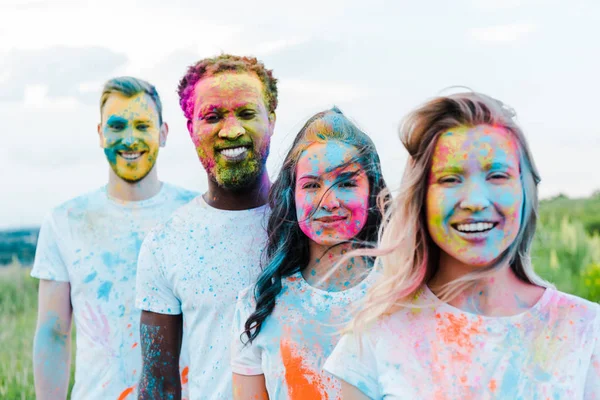 Enfoque selectivo de la mujer sonriendo cerca de amigos multiculturales con pinturas holi en las caras - foto de stock