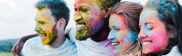 Plano panorámico de la mujer sonriendo cerca de amigos multiculturales con pinturas holi en las caras - foto de stock