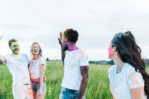 Foco seletivo de amigos multiculturais com tintas holi em rostos acenando as mãos — Fotografia de Stock