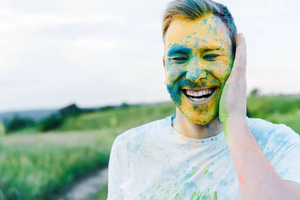 Happy young man with yellow and blue holi paints on face — Stock Photo