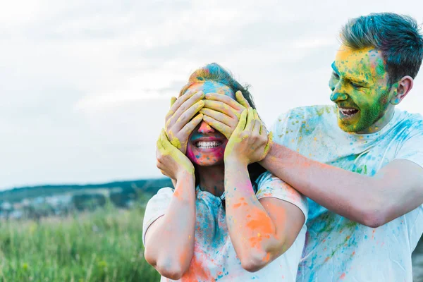 Jovem feliz sorrindo enquanto cobre o rosto da mulher com tintas holi no rosto — Fotografia de Stock