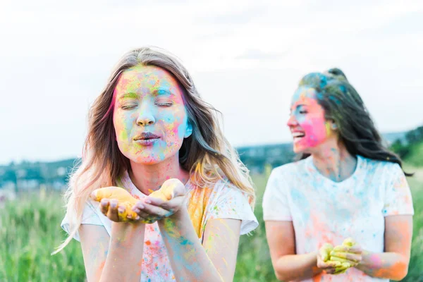 Messa a fuoco selettiva della donna con gli occhi chiusi soffiando polvere colorata vicino alla ragazza — Foto stock