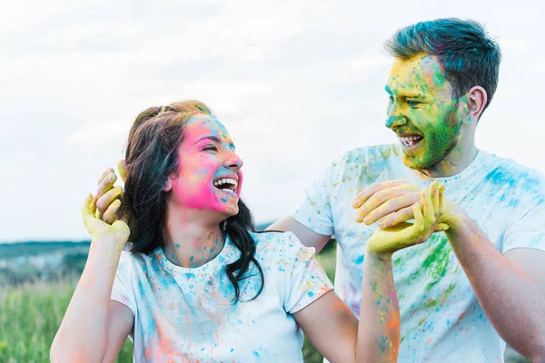 Feliz joven mujer sonriendo mientras gesticulando cerca de hombre con pinturas holi en la cara - foto de stock