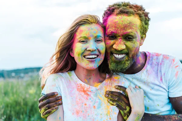 Happy african american man hugging cheerful young woman with holi paints on face — Stock Photo