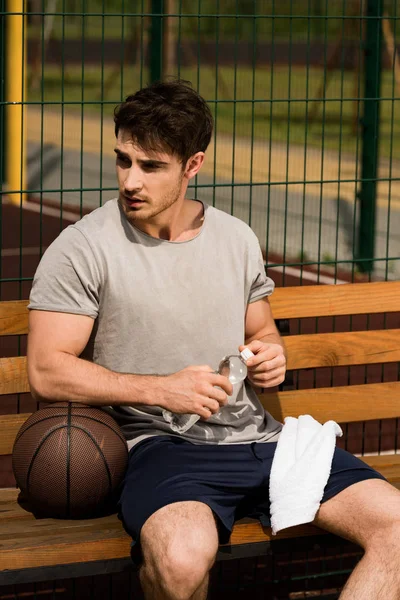 Basketballspieler sitzt auf Holzbank und hält Handtuch und Wasserflasche am Basketballplatz — Stockfoto