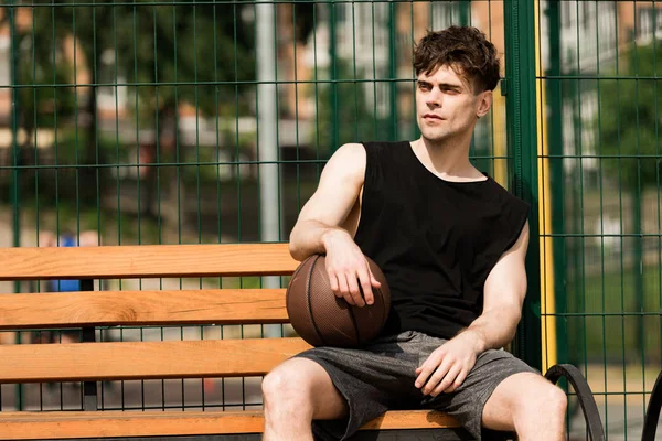 Seriöser Basketballspieler mit Ball sitzt bei sonnigem Tag auf Holzbank am Basketballplatz — Stockfoto