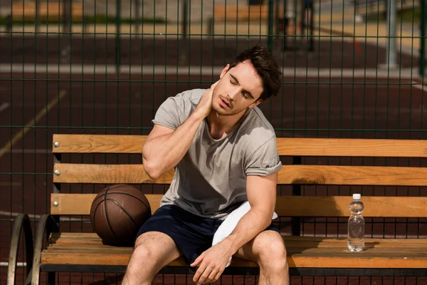 Jugador de baloncesto sentado en un banco de madera y sosteniendo la toalla y la botella de agua en la cancha de baloncesto - foto de stock