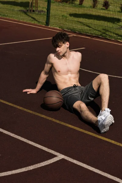Sexy jugador de baloncesto sin camisa con pelota sentado en la cancha de baloncesto - foto de stock