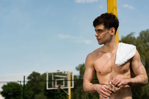 Sexy shirtless sportsman with towel opening bottle of water at basketball court — Stock Photo