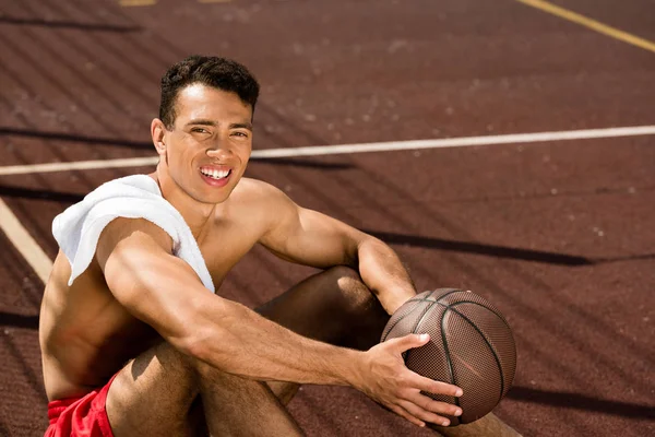 Shirtless mixed race basketball player smiling while sitting at basketball court with ball in sunny day — Stock Photo