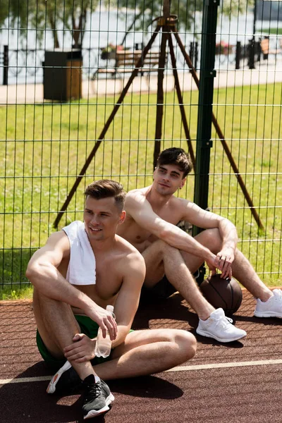 Two smiling shirtless basketball playes sitting at basketball court in sunny day — Stock Photo