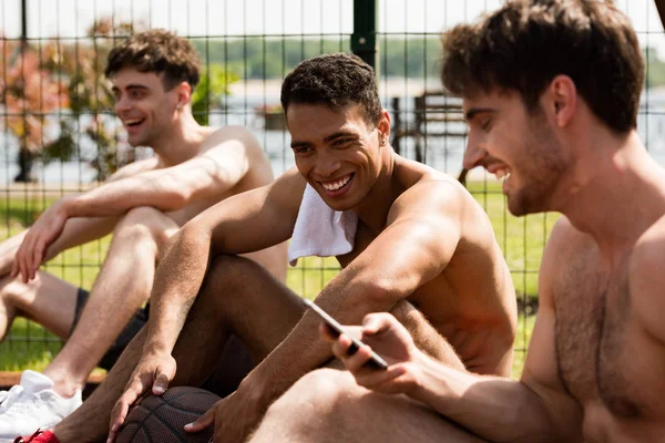 Three smiling shirtless basketball players with ball sitting at basketball court in sunny day — Stock Photo