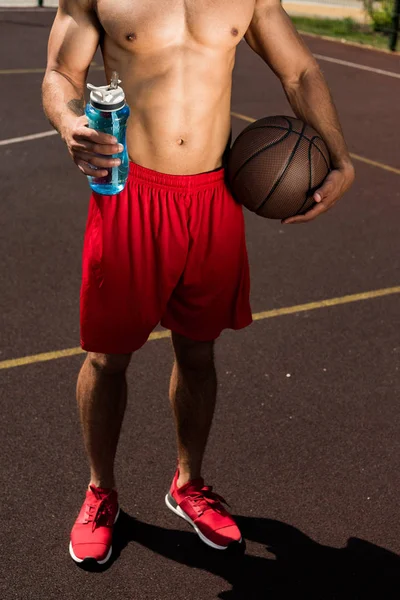 Visão parcial de desportista muscular sem camisa segurando garrafa de esporte e bola na quadra de basquete — Fotografia de Stock