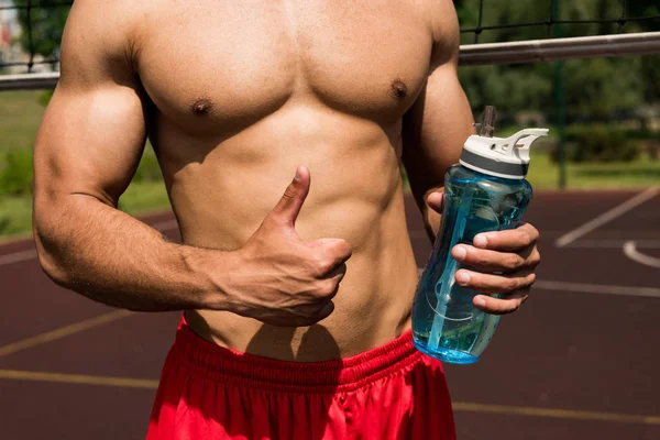 Partial view of shirtless muscular sportsman holding sport bottle and showing thumb up at basketball court — Stock Photo